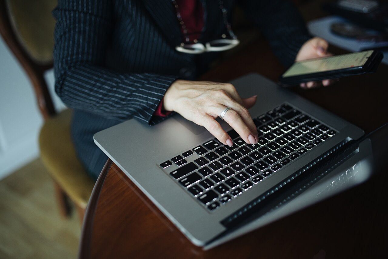 Middle aged woman working on laptop while holding smartphone at a wooden table