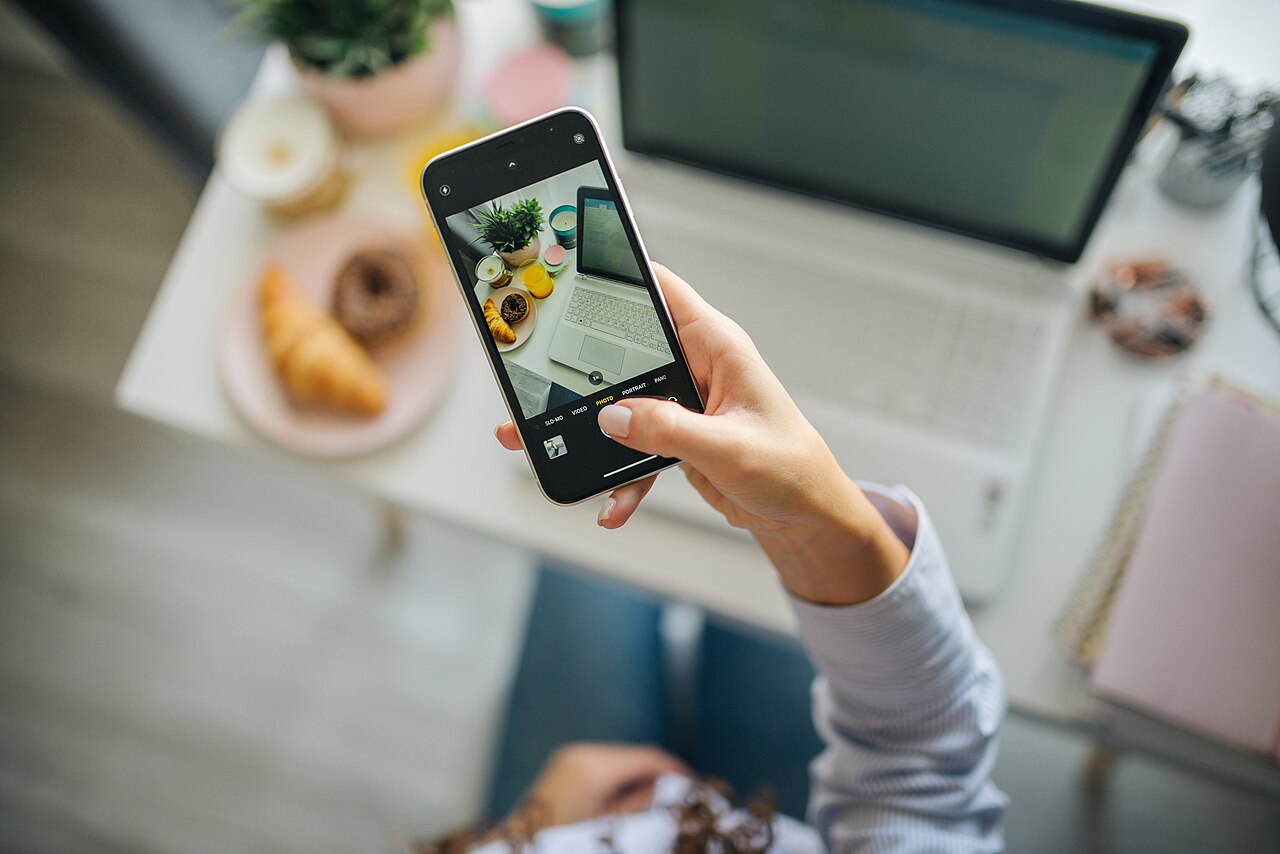 Person capturing a stylish workspace with smartphone and delicious snacks