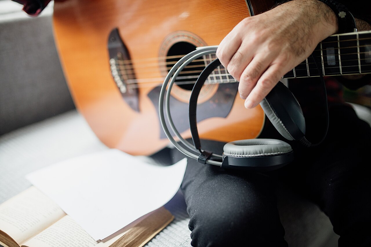 Person holds headphones while playing acoustic guitar and has sheet music on the table at home